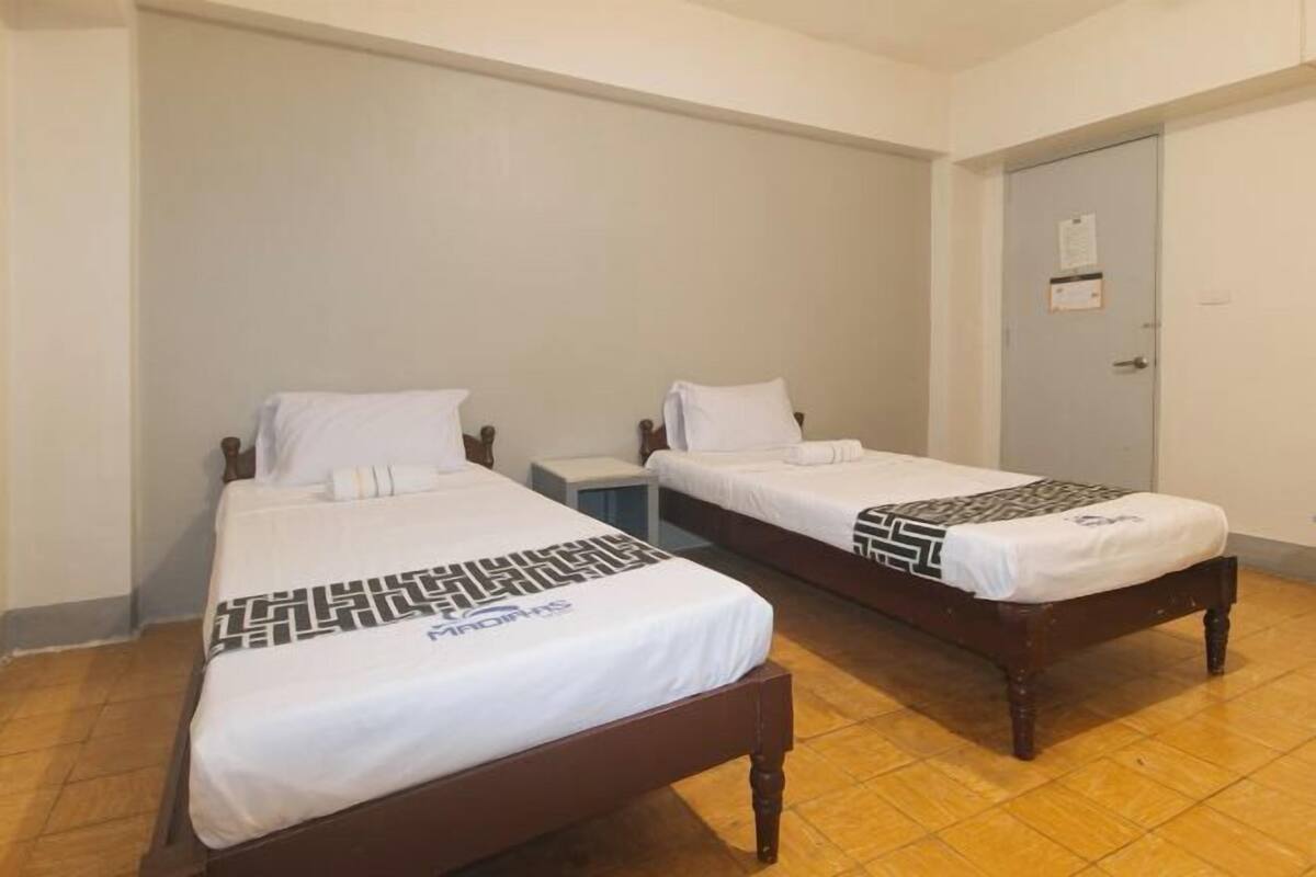 Una habitación con dos camas individuales, cabeceros de madera, ropa de cama blanca, una mesita de noche pequeña y una puerta con un aviso.