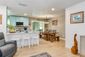 A modern kitchen with teal cabinets, a dining area with a wooden table and chairs, and a living room with a gray sofa and a wooden sculpture.