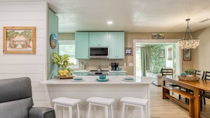 Une cuisine moderne avec des meubles de couleur turquoise, un îlot central doté de tabourets, un coin repas avec une table en bois et un salon avec un canapé gris.