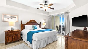A hotel room with a large bed, wooden headboard, two bedside tables with lamps, a flat-screen TV, a balcony with a table and chairs, and a ceiling fan. - The Crescent 210 (Miramar Beach)