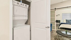 Un lavadero con lavadora y secadora Whirlpool, una puerta que da a un dormitorio con cama, una ventana con persianas y una mesita con bandeja.