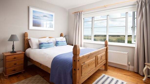 A bedroom with a wooden bed, a nightstand, a lamp, a window with curtains, and a view of the countryside. - Driftwood (Alnwick)