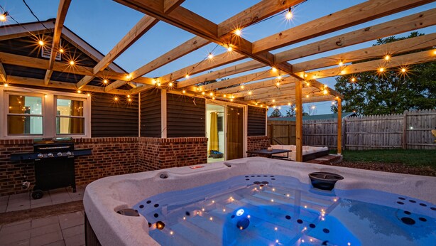 A hot tub under a wooden pergola with string lights, a barbecue grill, and a brick exterior wall.