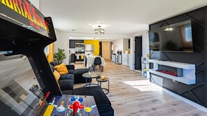A modern living room with a flat-screen TV, a gaming table, and a kitchen in the background.