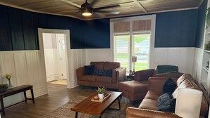 A living room with dark blue walls, a wooden ceiling, and a large window with blinds. There are two brown leather sofas, a wooden coffee table, and a small side table with a lamp.
