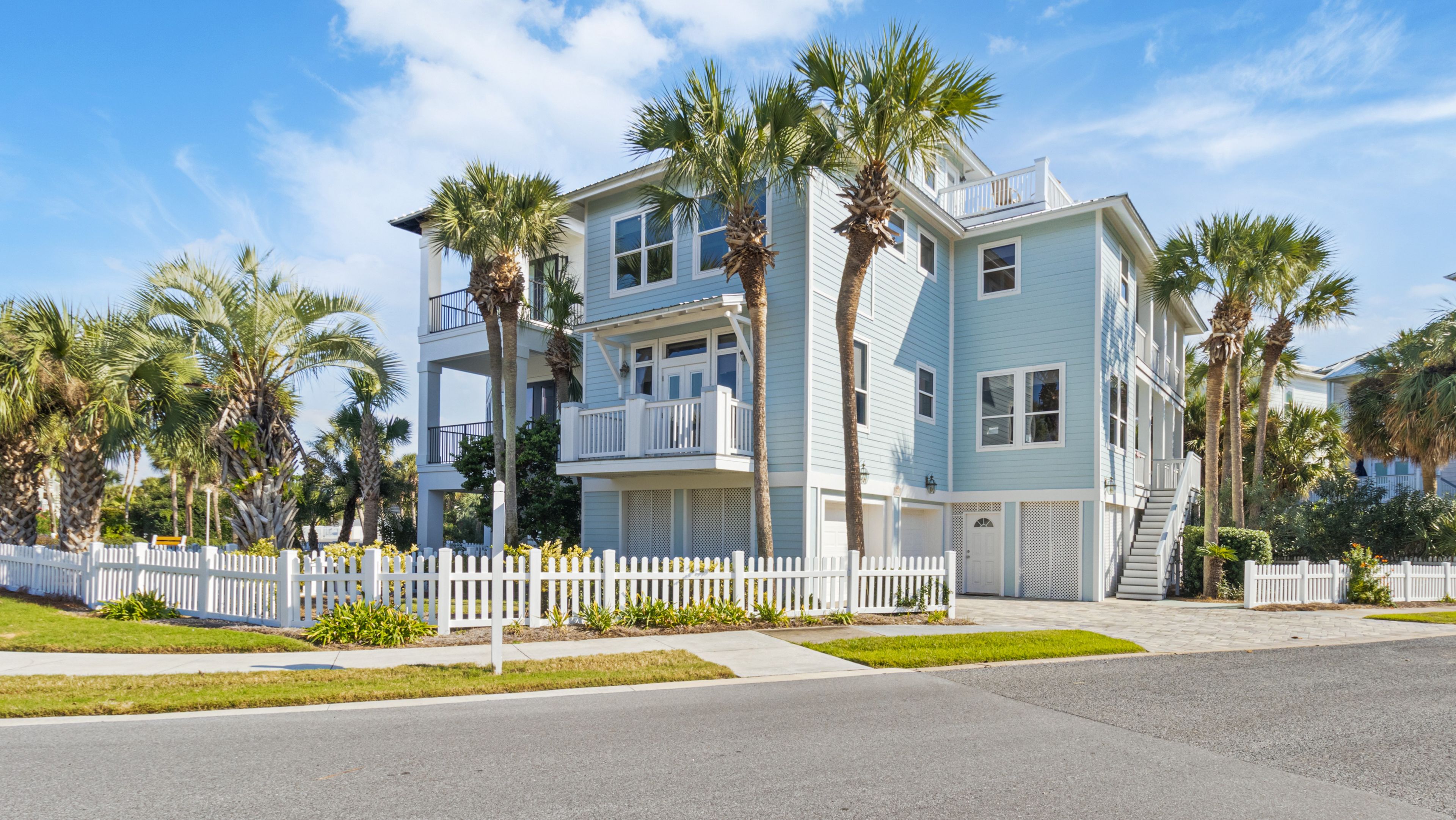 A two-story beachfront house with a white picket fence, palm trees, and a clear sky.