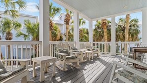 Een overdekt terras buiten met houten meubels, waaronder schommelstoelen en een bankje, met uitzicht op een woongebied met palmbomen en een huis.