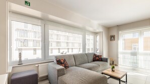 Un salon moderne avec un canapé d’angle gris, une petite table sur laquelle se trouve une bouteille et un banc.