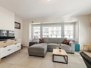 A modern living room with a grey sofa, a wooden coffee table, a flat-screen TV on a white cabinet, and large windows with a city view.