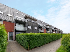 A row of modern apartment buildings with balconies, a paved walkway, and neatly trimmed hedges.