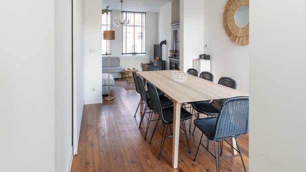 Un espace de repas moderne avec une table en bois et des chaises noires, un lustre, et une vue sur une cuisine et un salon en arrière-plan.