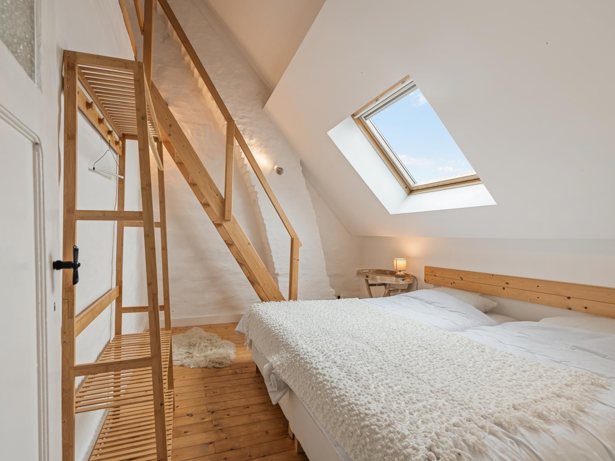A bedroom with a sloped ceiling, a skylight, a wooden staircase, a bed with white bedding, and a small bedside table with a lamp.