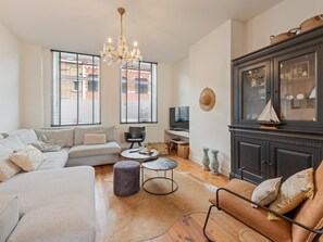 A spacious living room with a large sofa, a coffee table, and a fireplace. There are two windows with blinds, a chandelier, and a cabinet with decorative items. - Spacious town house in the centre of Nieuwpoort (Nieuwpoort)