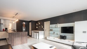 Una cucina moderna con isola centrale, una zona pranzo con tavolo bianco e sedie nere, e un soggiorno con TV e scaffali.