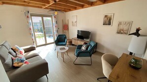 Una sala de estar con un sofá, un sillón y una mesa con una lámpara. Hay un televisor sobre un mueble bajo y una ventana con cortinas.