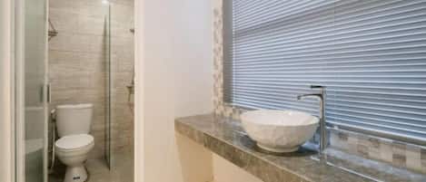 Un baño con cabina de ducha de vidrio, inodoro blanco y encimera de mármol con lavamanos sobre ella.