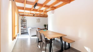 Uma área de refeições moderna com mesa e cadeiras de madeira, um sofá branco e uma estante de livros ao fundo.