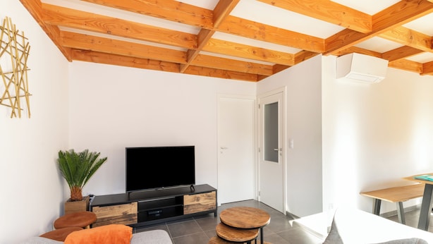 Uma sala de estar moderna com teto de madeira, uma TV de tela plana sobre um móvel de madeira, um sofá cinza com almofadas laranja e uma pequena mesa de madeira.