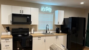 A modern kitchen with white cabinets, a black microwave, a stove, and a refrigerator. There is a neon sign that says "Let's Party" above the window. - Bring Kids & the Dog! House, Fence + Bunkhouse (Port Charlotte)