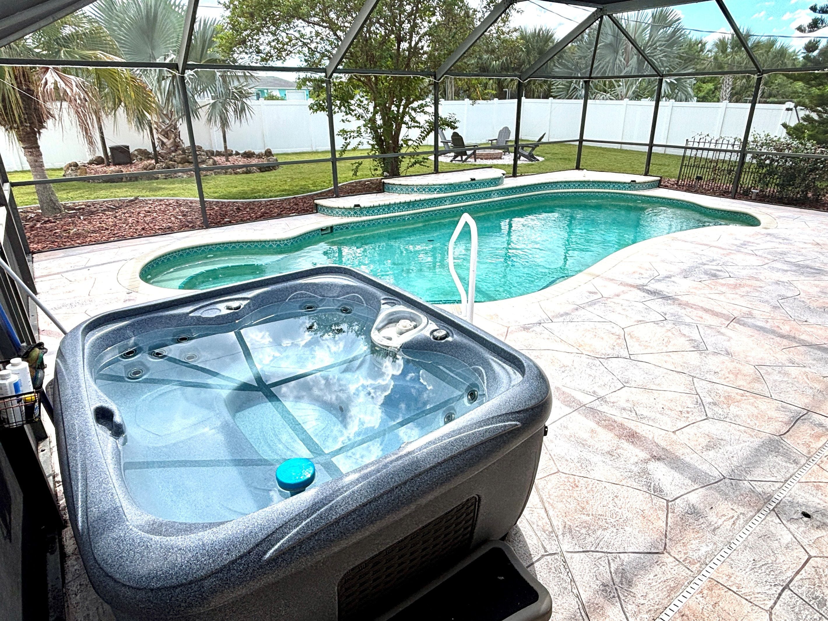 A hot tub in a backyard with a swimming pool, surrounded by a fence and palm trees.