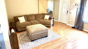 A living room with a sectional sofa, an ottoman, a patterned rug, a ceiling fan, and a door with a decorative glass panel. - Bring Kids & the Dog! House, Fence + Bunkhouse (Port Charlotte)