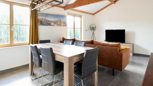 A modern living room with a wooden ceiling, a dining table with chairs, a sofa, and a TV.