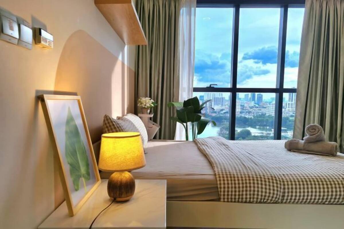 A hotel room with a bed, a bedside lamp, a framed picture, a plant, and a view of a cityscape through large windows.