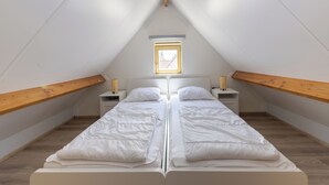 Une petite chambre mansardée, aux murs blancs, avec deux lits, des poutres apparentes et un puits de lumière.