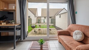 A living room with a sliding glass door leading to a patio, a sofa, a TV, and a small table with a plant. - Cosy holiday home on the Westhinder domain (Koksijde)