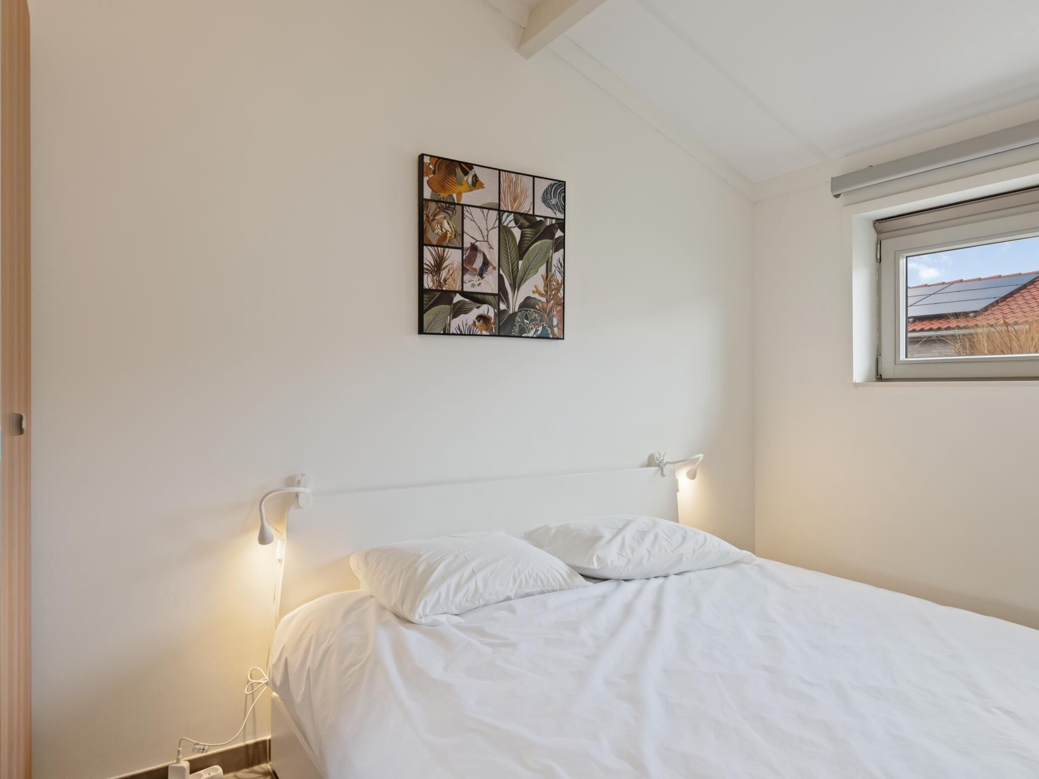 Et soveværelse med seng, et billede på væggen, en sengetavlelampe og et vindue med udsigt til en bygning.