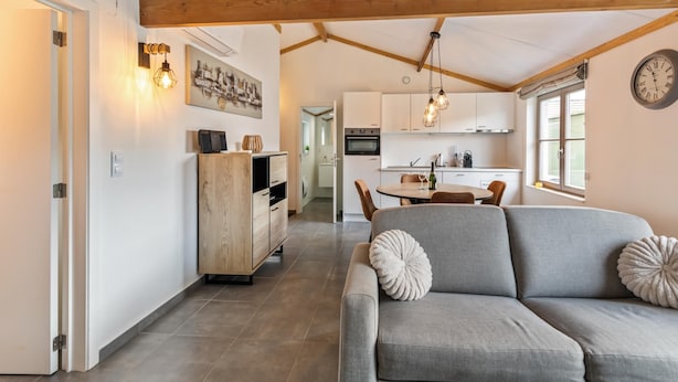 A modern kitchen with wooden beams, a dining area with a grey sofa, and a hallway leading to a bathroom. - Pet-friendly holiday home by the coast (Koksijde)