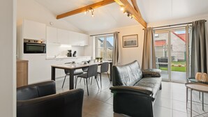 A modern kitchen with white cabinets, a dining table with chairs, a leather sofa, and a view of the outdoors through large windows. - Holiday home on park, pets allowed (Koksijde)