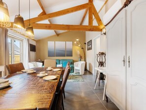 Un área de comedor con una mesa de madera, sillas e iluminación colgante. Hay un sofá y una mesita con un jarrón.