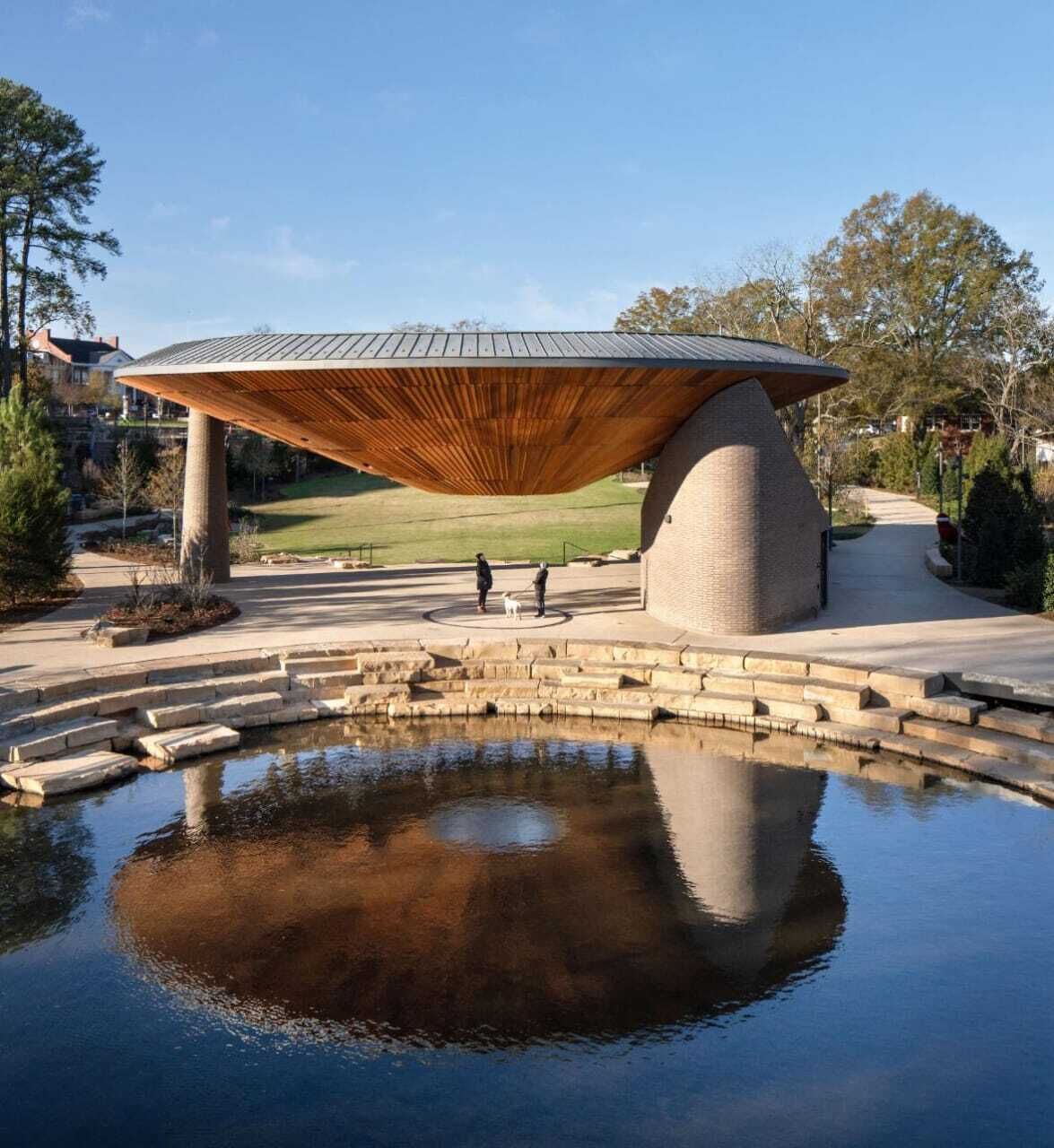 A modern pavilion with a unique roof structure, a central water feature, and a surrounding paved area.
