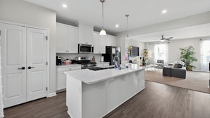 A modern kitchen with white cabinetry, a central island, stainless steel appliances, pendant lighting, and a view to a living area with a sofa and a plant. - 
Lux Home - Mins to Downtown, RDU, Fenton, UNC Health, WakeMed, & The Triangle (Cary)
