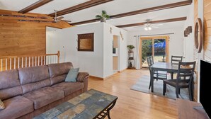 Een ruime woonkamer met een bruine leren bank, een eettafel met stoelen, houten balken in het plafond en uitzicht naar buiten.