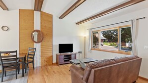 Een woonkamer met een bruine leren bank, een houten eettafel met stoelen, een televisie op een houten kast, en een groot raam met uitzicht.