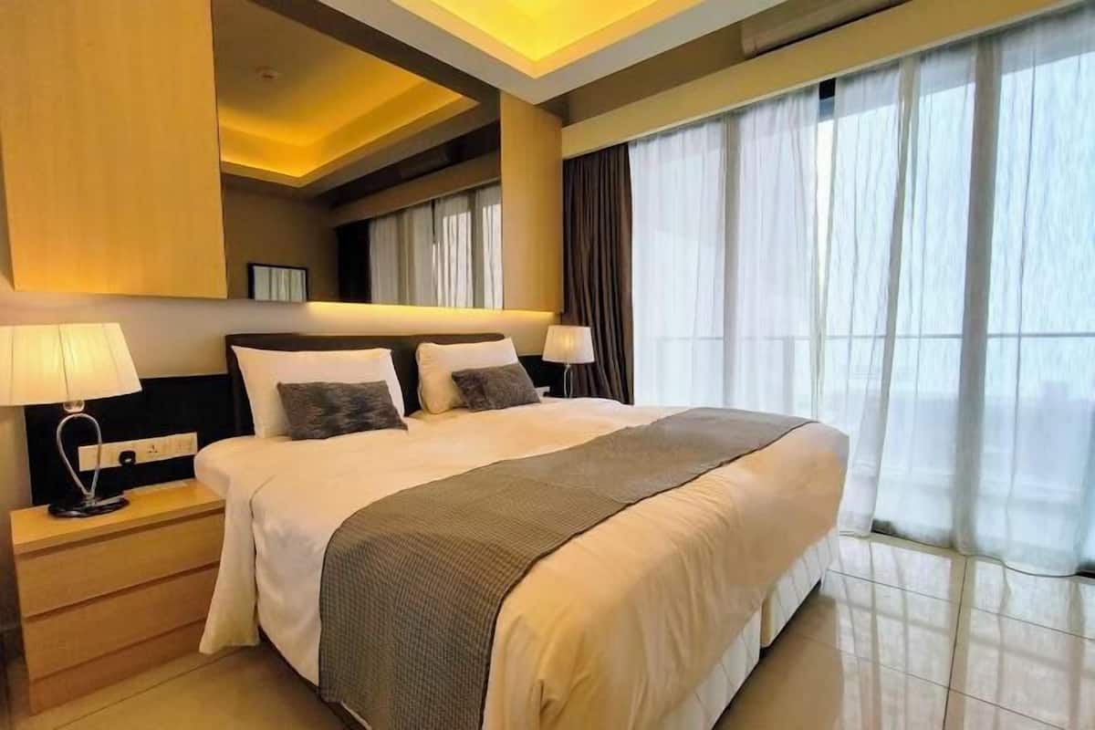 A modern hotel room with a large bed, two bedside tables with lamps, a headboard, and a large window with curtains.