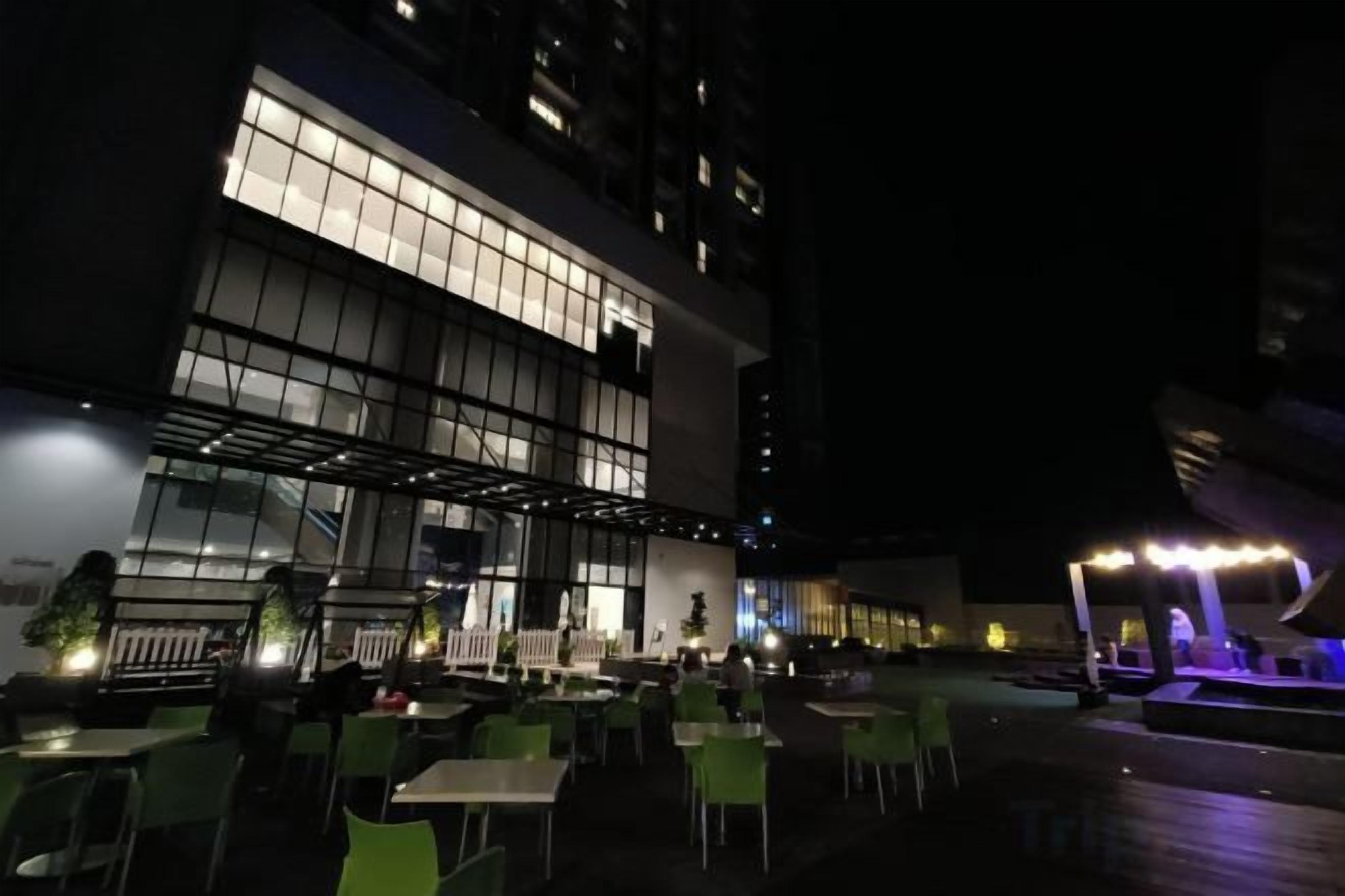 An outdoor dining area with tables and chairs, a building with large windows, and a well-lit entrance.