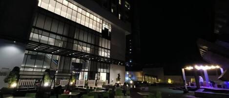 An outdoor dining area with tables and chairs, a building with large windows, and a well-lit entrance.