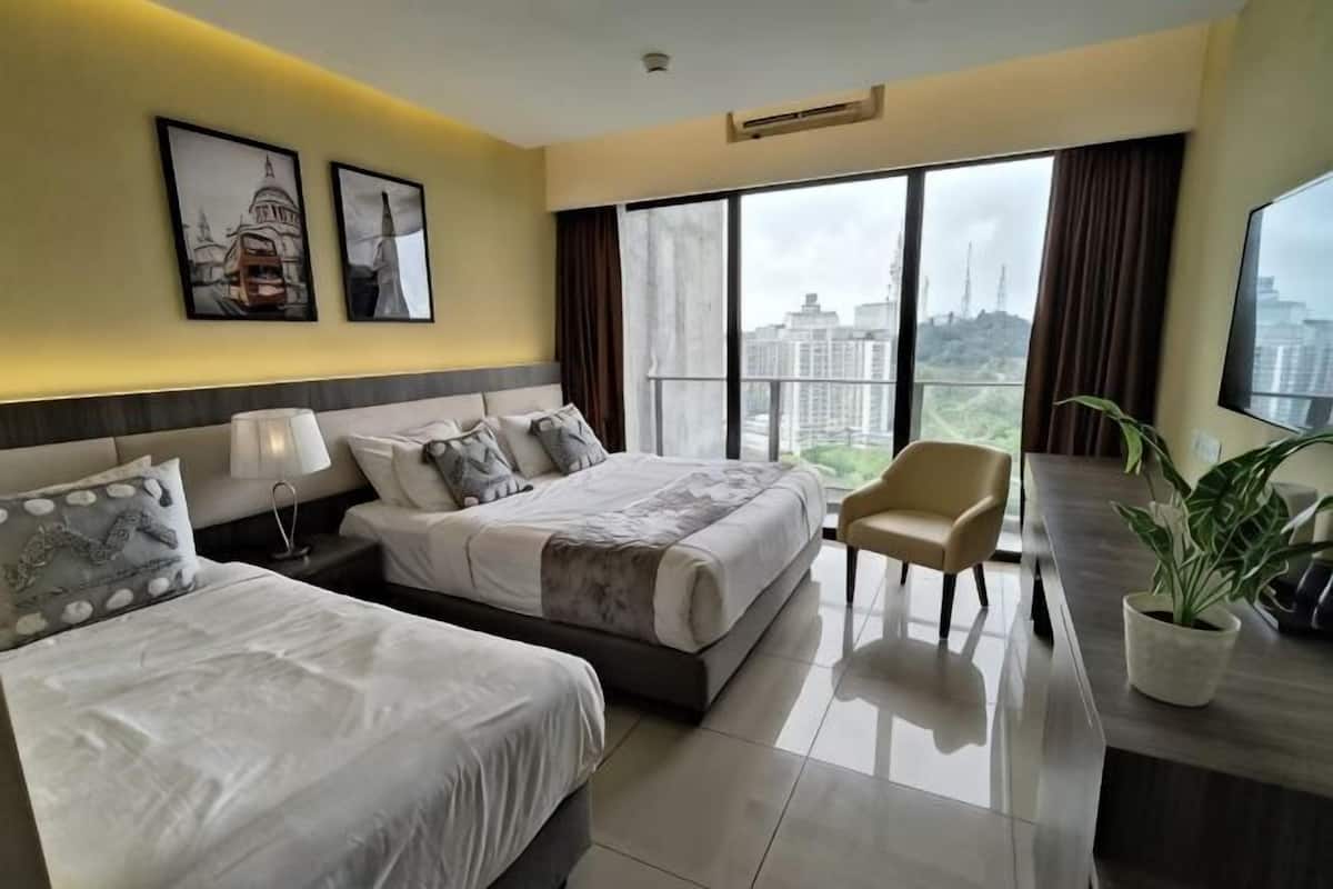A hotel room with two beds, a desk, a chair, a plant, and large windows with a view.