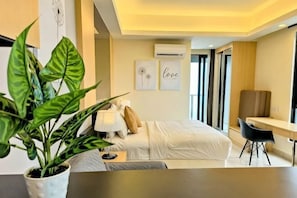 A modern hotel room with a bed, bedside table, lamp, potted plant, and a desk with a chair.