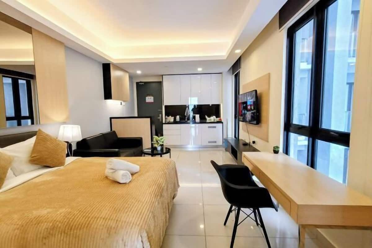 A modern hotel room with a bed, a desk, a chair, a TV, and a kitchenette in the background.