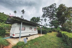 A white RV parked in a lush garden with a covered porch, surrounded by greenery and trees. - Samui Camping Farm (Koh Samui)