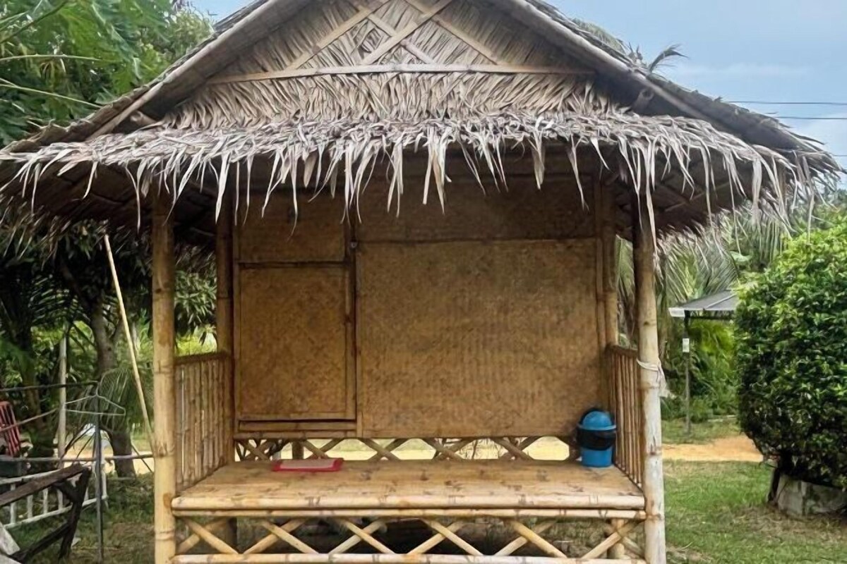 A thatched-roof hut with a wooden frame, a bench, and a blue barrel.