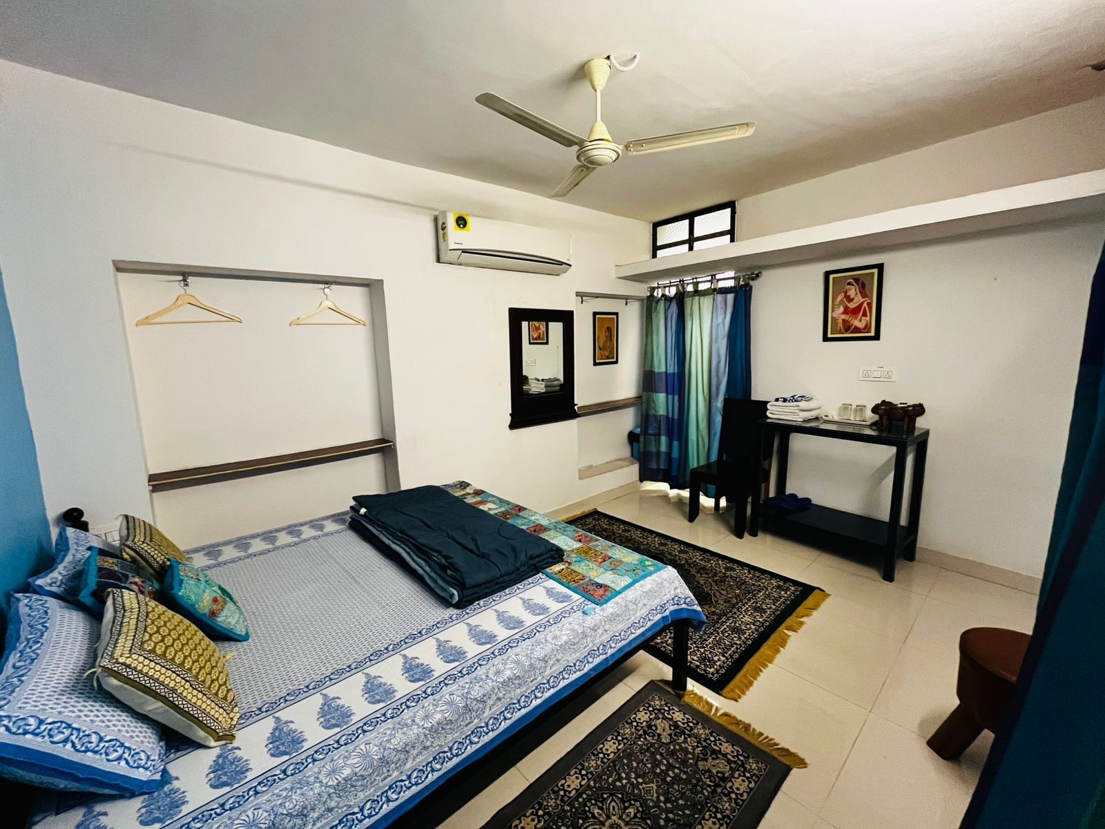 A compact room with a bed, a desk, and a chair. There is a ceiling fan, an air conditioning unit, and a window with curtains.