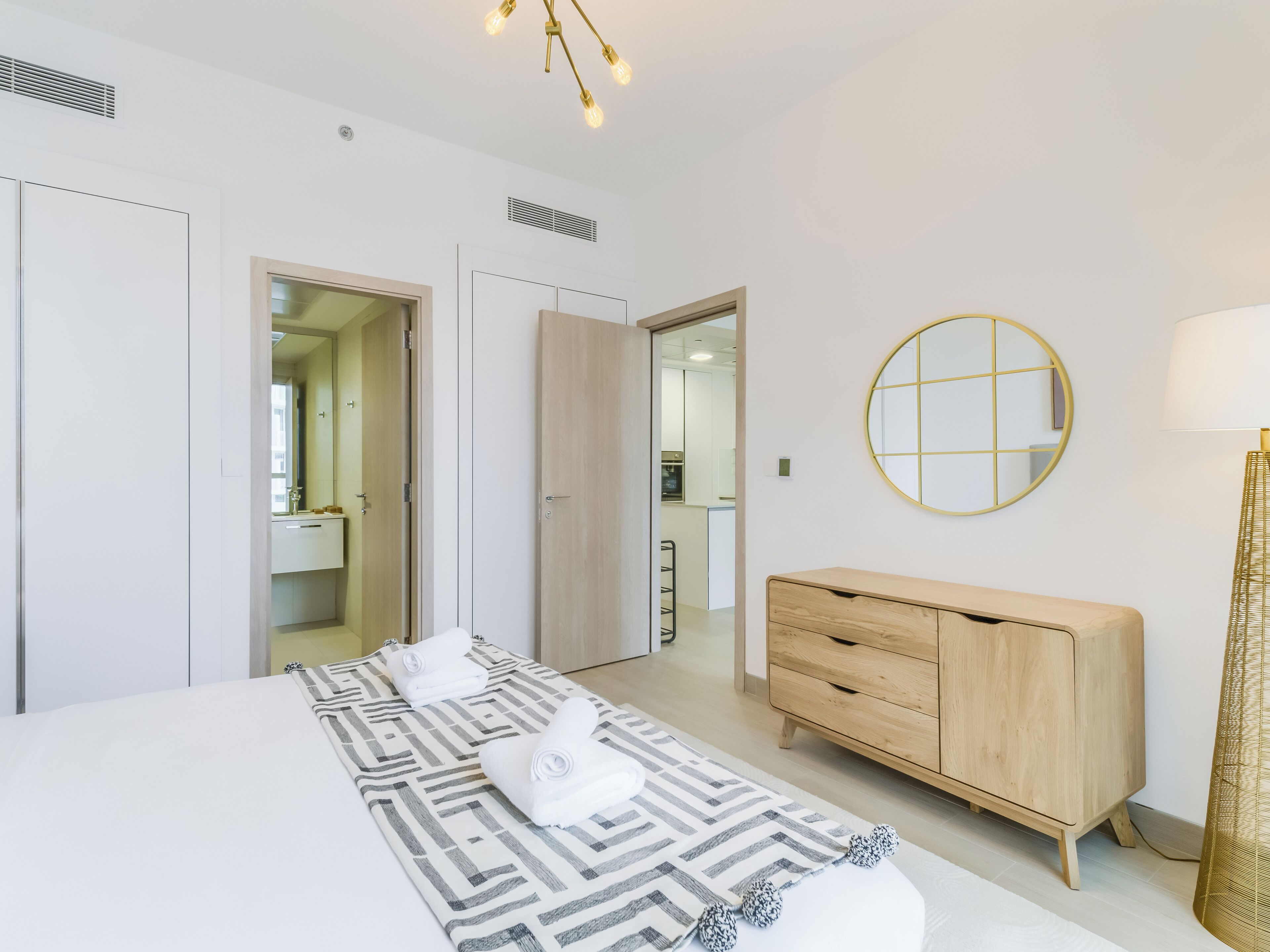 A modern hotel room with a large bed, a wooden dresser, a round mirror, and a ceiling light fixture.
