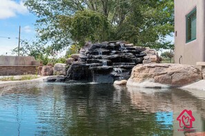 Ein Felswasserfall in einer angelegten Grünanlage mit Pool und einem Gebäude im Hintergrund.