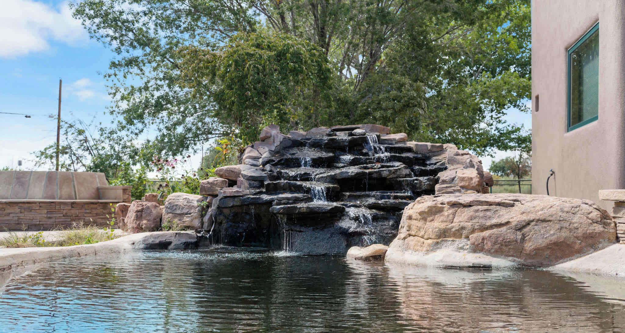 Une cascade artificielle en roche, avec un bassin à sa base, entourée de gros rochers et d’arbres.