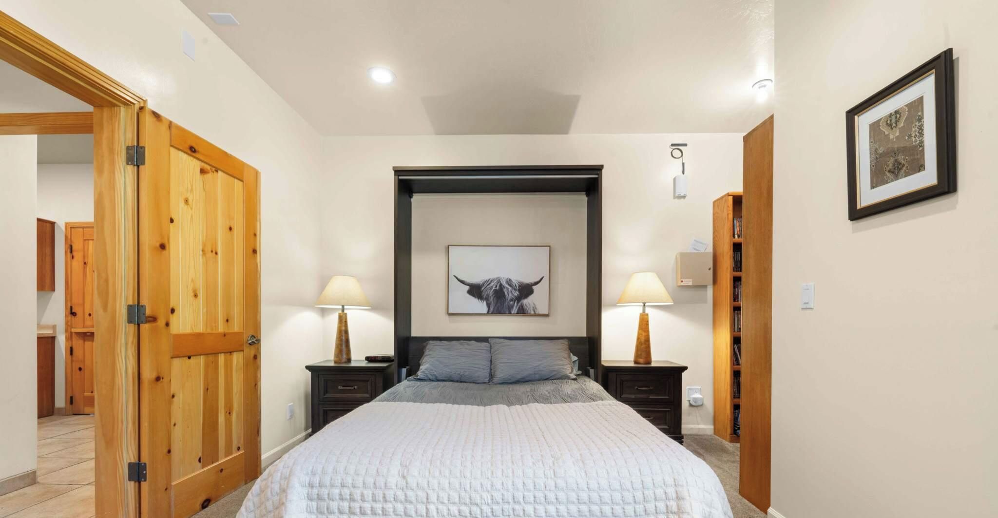 Une chambre à coucher avec un lit, deux tables de chevet avec des lampes, une armoire en bois, un tableau encadré au mur et une porte donnant sur une autre pièce.